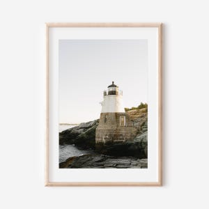 May include: A framed print of a coastal lighthouse. The lighthouse is white and black, set on a rocky cliffside. The ocean is visible in the background under a clear sky. The print is in a light wood frame.