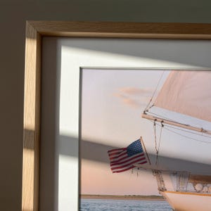 K&ouml;nnte beinhalten: Gerahmter Druck eines Segelboots mit amerikanischer Flagge. Das Bild zeigt ein wei&szlig;es Segelboot mit beigem Segel vor einem pastellfarbenen Himmel. Der Rahmen ist helles Holz mit wei&szlig;em Passepartout.