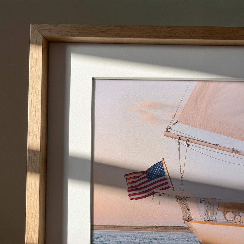K&ouml;nnte beinhalten: Gerahmter Druck eines Segelboots mit amerikanischer Flagge. Das Bild zeigt ein wei&szlig;es Segelboot mit beigem Segel vor einem pastellfarbenen Himmel. Der Rahmen ist helles Holz mit wei&szlig;em Passepartout.
