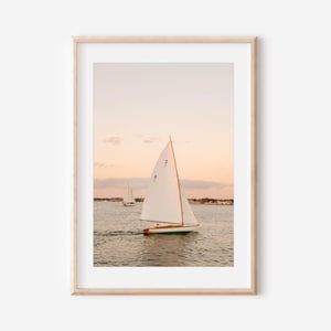 May include: A framed print featuring a sailboat with a white sail gliding across the water. The boat has a wooden mast and a green hull. The sky is a soft peach color, and another sailboat is visible in the distance.