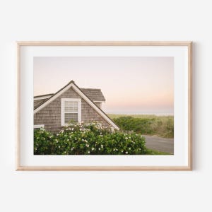Puede incluir: Una impresión enmarcada que presenta una escena costera. La imagen muestra una casa con un tejado de tejas grises y una ventana con marco blanco, rodeada de arbustos verdes con flores blancas. Un camino conduce al mar bajo un cielo suavemente rosado y amarillo.
