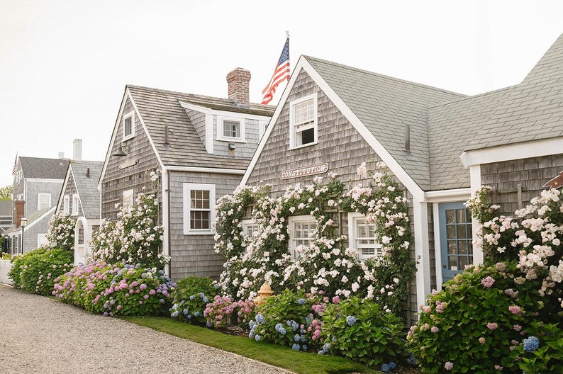 May include: A row of charming gray-shingled buildings with white trim and multiple windows. The buildings are adorned with climbing white and pink flowers and surrounded by lush green and colorful hydrangea bushes. An American flag waves in the sky.