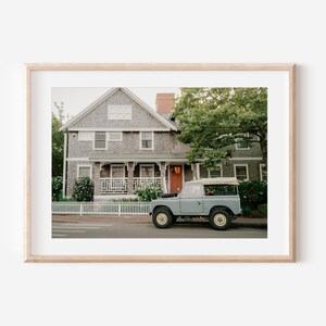 Op de afbeelding: Een lichtblauwe vintage Land Rover geparkeerd voor een grijs geschilderd huis met een wit hek. Het huis heeft een veranda met witte leuningen en een rode deur. Er zijn groene bomen en struiken op de achtergrond.