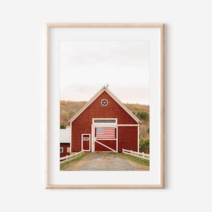 Puede incluir: Impresión enmarcada de un granero rojo con una bandera estadounidense en su interior. El granero tiene una puerta blanca y una valla blanca. Un camino de grava conduce al granero. El cielo es de un color suave y apagado.