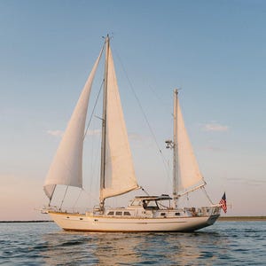 May include: A sailboat with white sails glides across the water under a blue sky. The boat has a white hull and a small American flag at the stern. The scene is bathed in the warm light of the setting sun.