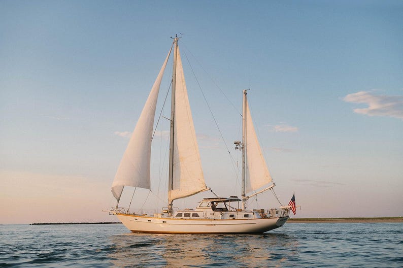 May include: A sailboat with white sails glides across the water under a blue sky. The boat has a white hull and a small American flag at the stern. The scene is bathed in the warm light of the setting sun.