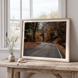 May include: A framed landscape photograph of a winding asphalt road flanked by trees with autumn foliage in shades of orange, yellow, and brown. The print is displayed in a light wood frame, resting on a rustic wooden table near a window.