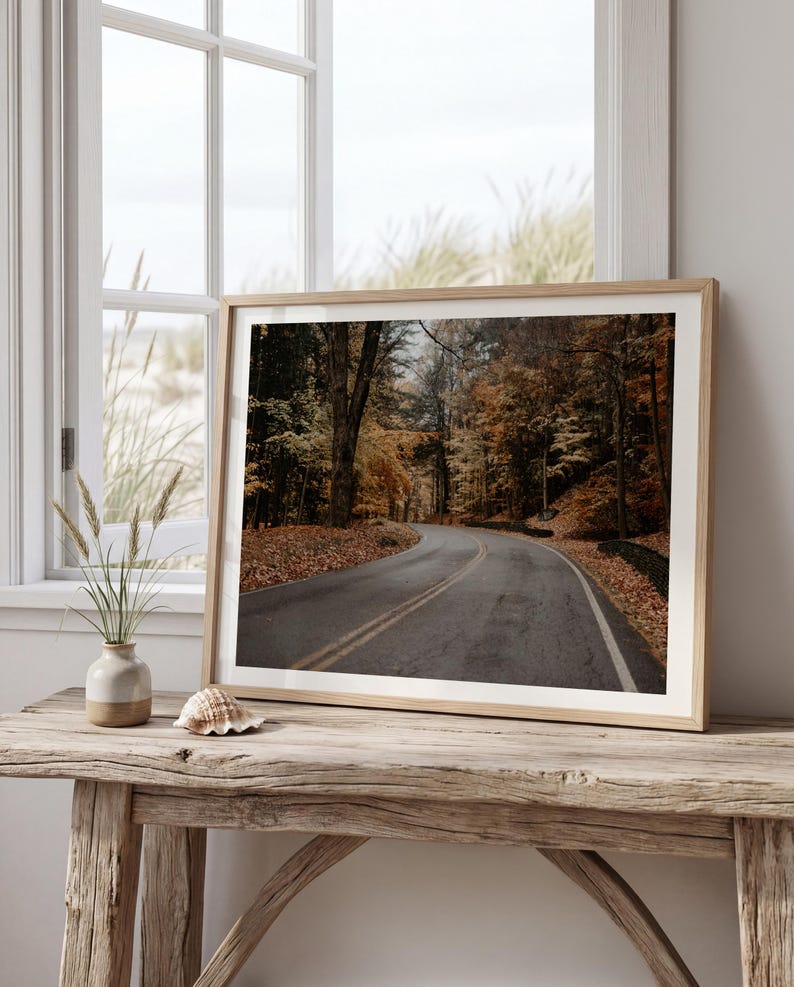 May include: A framed landscape photograph of a winding asphalt road flanked by trees with autumn foliage in shades of orange, yellow, and brown. The print is displayed in a light wood frame, resting on a rustic wooden table near a window.