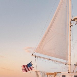 K&ouml;nnte beinhalten: Ein wei&szlig;es Segelboot mit einem gro&szlig;en wei&szlig;en Segel und einer amerikanischen Flagge am Bug. Das Boot befindet sich auf dem Wasser mit einem klaren Himmel im Hintergrund. Das Bild wurde bei Sonnenuntergang aufgenommen.