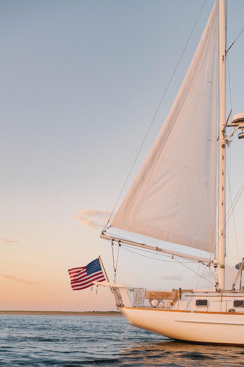 K&ouml;nnte beinhalten: Ein wei&szlig;es Segelboot mit einem gro&szlig;en wei&szlig;en Segel und einer amerikanischen Flagge am Bug. Das Boot befindet sich auf dem Wasser mit einem klaren Himmel im Hintergrund. Das Bild wurde bei Sonnenuntergang aufgenommen.
