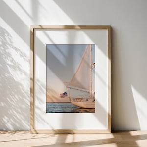 K&ouml;nnte beinhalten: Gerahmter Druck eines Segelbootes auf dem Wasser bei Sonnenuntergang. Das Boot ist wei&szlig; mit einem gro&szlig;en Segel und einer amerikanischen Flagge. Der Himmel ist eine Mischung aus Orange, Blau und Gelb. Der Rahmen ist hellbraun.