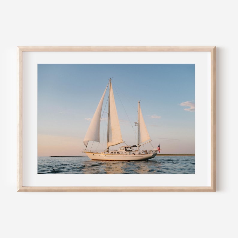 May include: A framed print of a sailboat on the water. The white sailboat has two masts with white sails. An American flag flies from the back of the boat. The sky is blue with a hint of pink and orange.