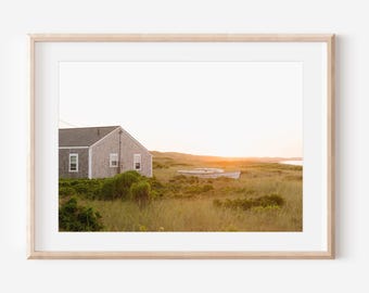 May include: A framed print of a coastal scene. A grey-shingled house with white trim sits on a grassy dune. A small boat rests in the tall grass. The sky is a warm orange and yellow.