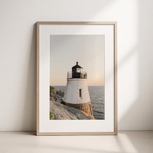 May include: A framed vertical wall art print featuring a white stone lighthouse with a black lantern room, situated on a rocky coastline overlooking the ocean at sunset. The frame is light wood with a white mat border surrounding the photograph.