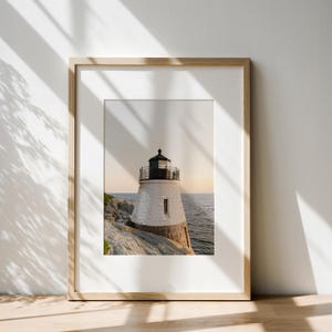 May include: A framed wall art print featuring a white stone lighthouse with a black lantern room, situated on a rocky coastline overlooking the ocean at sunset. The artwork is encased in a light-toned wooden frame with a white mat border.