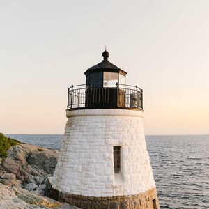 May include: A stone lighthouse featuring a white-painted upper section and a natural grey stone base. The structure includes a black metal lantern room with a railing, a single narrow window, and a dark roof, positioned on a rocky coastal cliff.