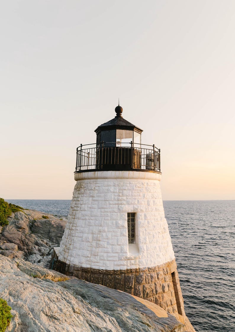May include: A stone lighthouse featuring a white-painted upper section and a natural grey stone base. The structure includes a black metal lantern room with a railing, a single narrow window, and a dark roof, positioned on a rocky coastal cliff.