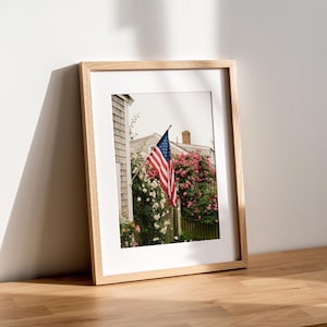 May include: A framed photograph featuring an American flag hanging from the side of a shingled house, surrounded by vibrant pink climbing roses and green foliage, with a chimney visible against a bright sky in the background.