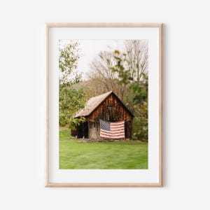 Puede incluir: Impresión enmarcada que muestra un granero de madera rústica con un tejado de metal rojo. Una bandera estadounidense cuelga de la fachada del granero. El granero está situado en un campo verde, rodeado de árboles, creando un paisaje rural.