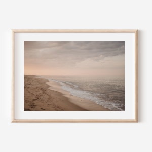 Op de afbeelding: Een foto van een zandstrand met een zachte golf die tegen de kust aan spoelt. De lucht is zacht roze en grijs, met wolken die schaduwen op het water werpen.