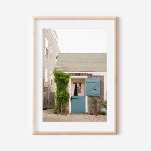 Puede incluir: Una impresión enmarcada de un edificio pintoresco con una puerta y contraventanas azules. El edificio tiene un letrero que dice "MARY SLADE". Una vid verde crece por el costado. La imagen está ambientada en un pueblo costero.