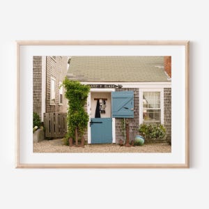 Puede incluir: Impresión enmarcada de una escena costera con un edificio desgastado, una puerta y una contraventana azules. El letrero sobre la puerta dice "MARY & SLADE". Anclas decorativas y una vid verde completan el tema náutico.