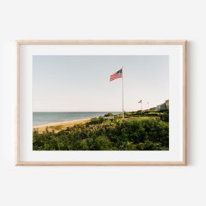 Könnte beinhalten: Ein Foto von einer Strandszene mit einer US-Flagge, die im Wind weht. Die Flagge befindet sich auf einem Mast vor einer Reihe von Häusern. Der Strand ist sandig und das Wasser ist blau. Der Himmel ist hellblau mit einigen Wolken.