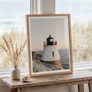 May include: A framed wall art print featuring a photograph of a white stone lighthouse with a black lantern room, situated on a rocky coastline overlooking the ocean at sunset, enclosed in a light-toned wooden frame with a white mat border.