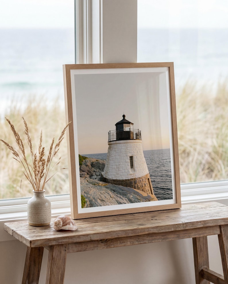 May include: A framed wall art print featuring a photograph of a white stone lighthouse with a black lantern room, situated on a rocky coastline overlooking the ocean at sunset, enclosed in a light-toned wooden frame with a white mat border.