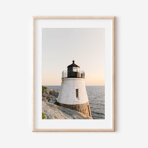 May include: A framed wall art print featuring a white stone lighthouse with a black lantern room and railing, situated on a rocky coastline overlooking the ocean during a soft sunset with a pale sky and calm water in the background.