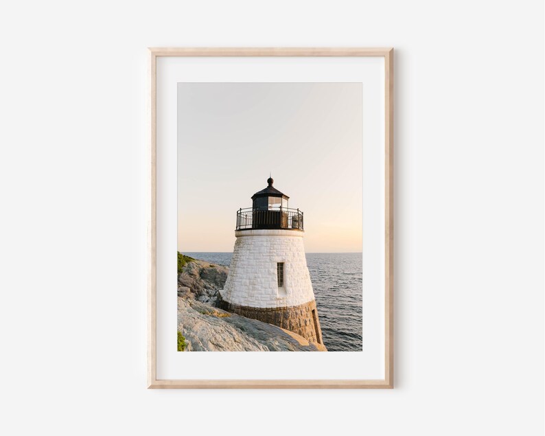 May include: A framed wall art print featuring a white stone lighthouse with a black lantern room and railing, situated on a rocky coastline overlooking the ocean during a soft sunset with a pale sky and calm water in the background.