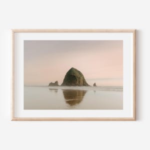 Op de afbeelding: Een ingelijste foto van een grote rotsformatie die uit de oceaan steekt. De rots is groen en bedekt met vegetatie. Het water is kalm en reflecteert de lucht, die zacht roze en oranje is.