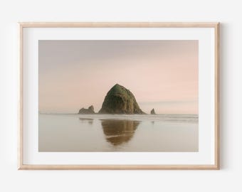 May include: A framed photograph of a large rock formation jutting out of the ocean. The rock is green and covered in vegetation. The water is calm and reflects the sky, which is a soft pink and orange.