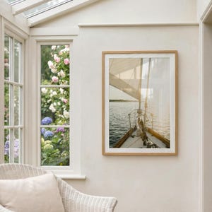 May include: A framed photograph of a sailboat sailing on the water. The image is framed in a light brown wooden frame. The sailboat's sail is up, and the sun is shining on the water. The photograph is hanging on a white wall.
