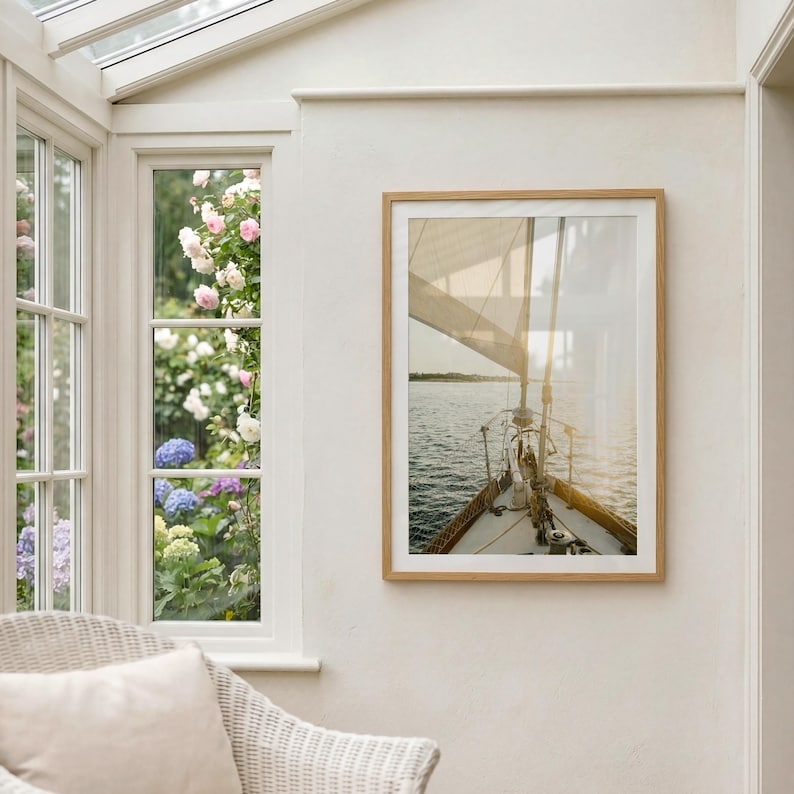 May include: A framed photograph of a sailboat sailing on the water. The image is framed in a light brown wooden frame. The sailboat's sail is up, and the sun is shining on the water. The photograph is hanging on a white wall.