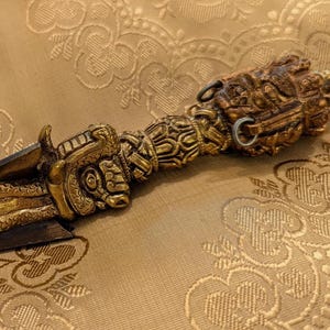 Peut inclure: Un poignard orné avec un manche de couleur bronze et une lame sombre. La poignée présente des sculptures complexes et un motif de serpent. Le poignard repose sur un tissu doré à motifs.