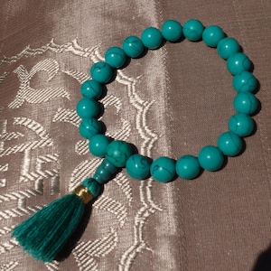 Könnte beinhalten: Ein türkisfarbenes Perlenarmband mit einem grünen Quastenanhänger. Das Armband besteht aus runden Perlen und hat ein schlichtes Design.