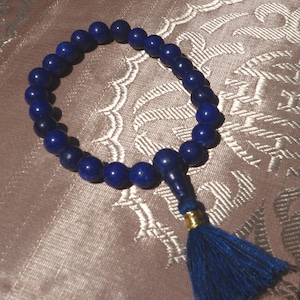 Könnte beinhalten: Ein blaues Lapislazuli-Perlenarmband mit Quaste. Die Perlen sind rund und haben eine dunkelblaue Farbe. Die Quaste ist aus blauem Stoff und hat einen goldenen Akzent.