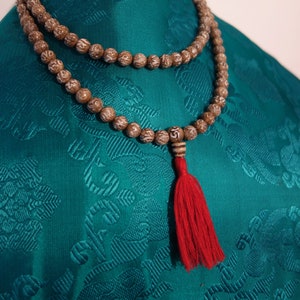 Könnte beinhalten: Eine braune Holz-Mala-Kette mit einem roten Quastenanhänger, die auf einem blaugrünen Stoffhintergrund liegt.