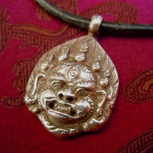 Può includere: Collana con ciondolo in argento che presenta una rappresentazione tridimensionale dettagliata e ornata del viso di una creatura mitologica con un terzo occhio. Il ciondolo è attaccato a un cordino in pelle nera.