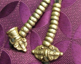 May include: Two ornate, gold-colored metal objects with beaded handles and leather straps, resting on a purple patterned fabric. The objects have detailed, decorative ends, suggesting a historical or cultural significance.