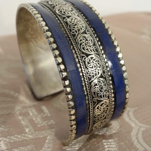 Op de afbeelding: Zilveren manchetarmband met ingewikkeld filigraan en donkerblauwe inleg. De armband heeft een brede band met gedetailleerde krullen en een rij kleine, zilveren kralen langs de randen. Een opvallend sieraad.