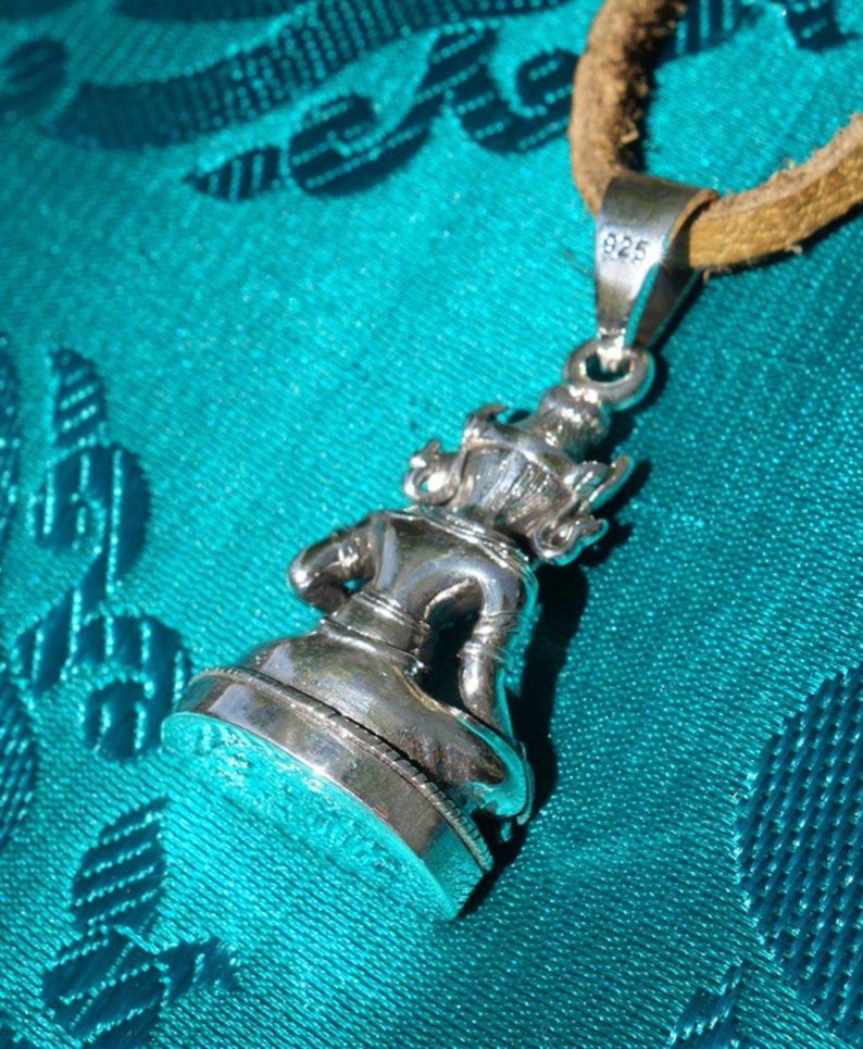 May include: Silver pendant featuring a detailed sculpture of a seated figure. The pendant is attached to a brown leather cord and has a 925 stamp on the back.