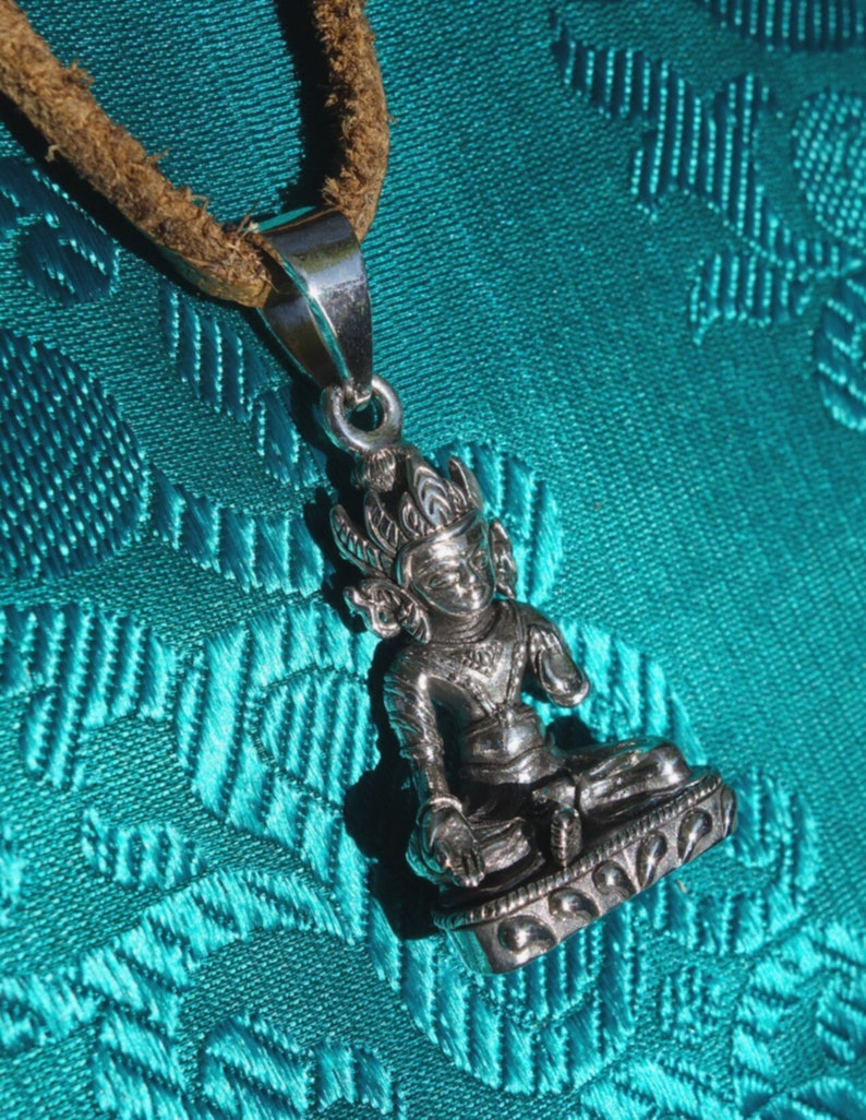 May include: Silver pendant necklace featuring a detailed depiction of a seated Buddha figure with a crown and intricate details. The pendant is on a brown leather cord and is displayed against a turquoise background.