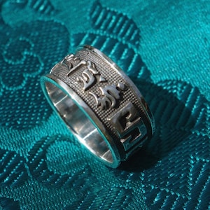 Könnte beinhalten: Ein silberner Ring mit einem drehbaren Band, das mit einem Mantra in tibetischer Schrift verziert ist. Der Ring liegt auf einem türkisfarbenen Stoffhintergrund.
