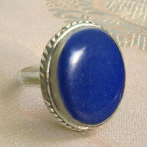 Op de afbeelding: Een zilveren ring met een grote, ovale, diepblauwe edelsteen. De edelsteen is gezet in een zilveren bezel met een gedraaid touwontwerp. De ringband is eenvoudig en zilver.