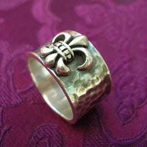 Könnte beinhalten: Ein silberner Ring mit einer strukturierten Band und einem Fleur-de-lis-Design.
