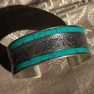 Peut inclure: Bracelet manchette en argent orné d'incrustations de pierres turquoise et de détails en filigrane. Le bracelet a une large bande avec des rectangles turquoise et une bande centrale décorative. Le design ouvert facilite le port.