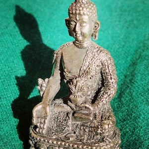 May include: A detailed bronze Buddha statue, seated in a meditative pose. The figure is adorned with intricate patterns on the robe and base. The statue is set against a green backdrop, highlighting the detailed craftsmanship of the religious sculpture.