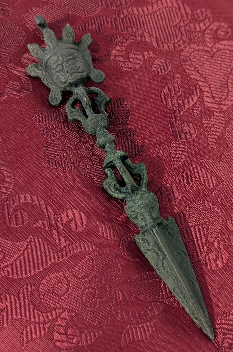 May include: A bronze-colored metal ritual dagger with a decorative handle featuring a stylized face and intricate designs. The dagger is on a red fabric background.